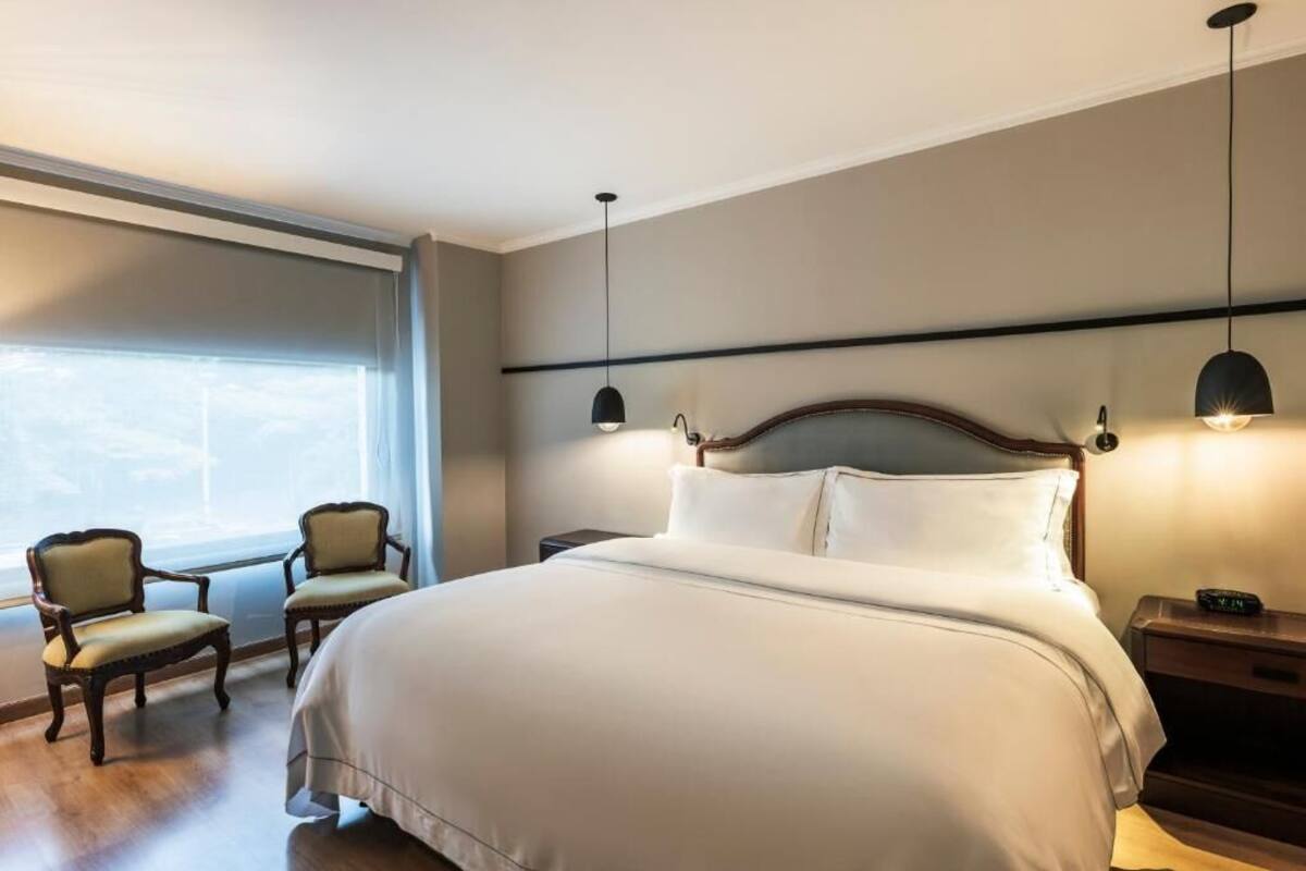 A hotel room with a large bed, two chairs, a nightstand, and a window with curtains.