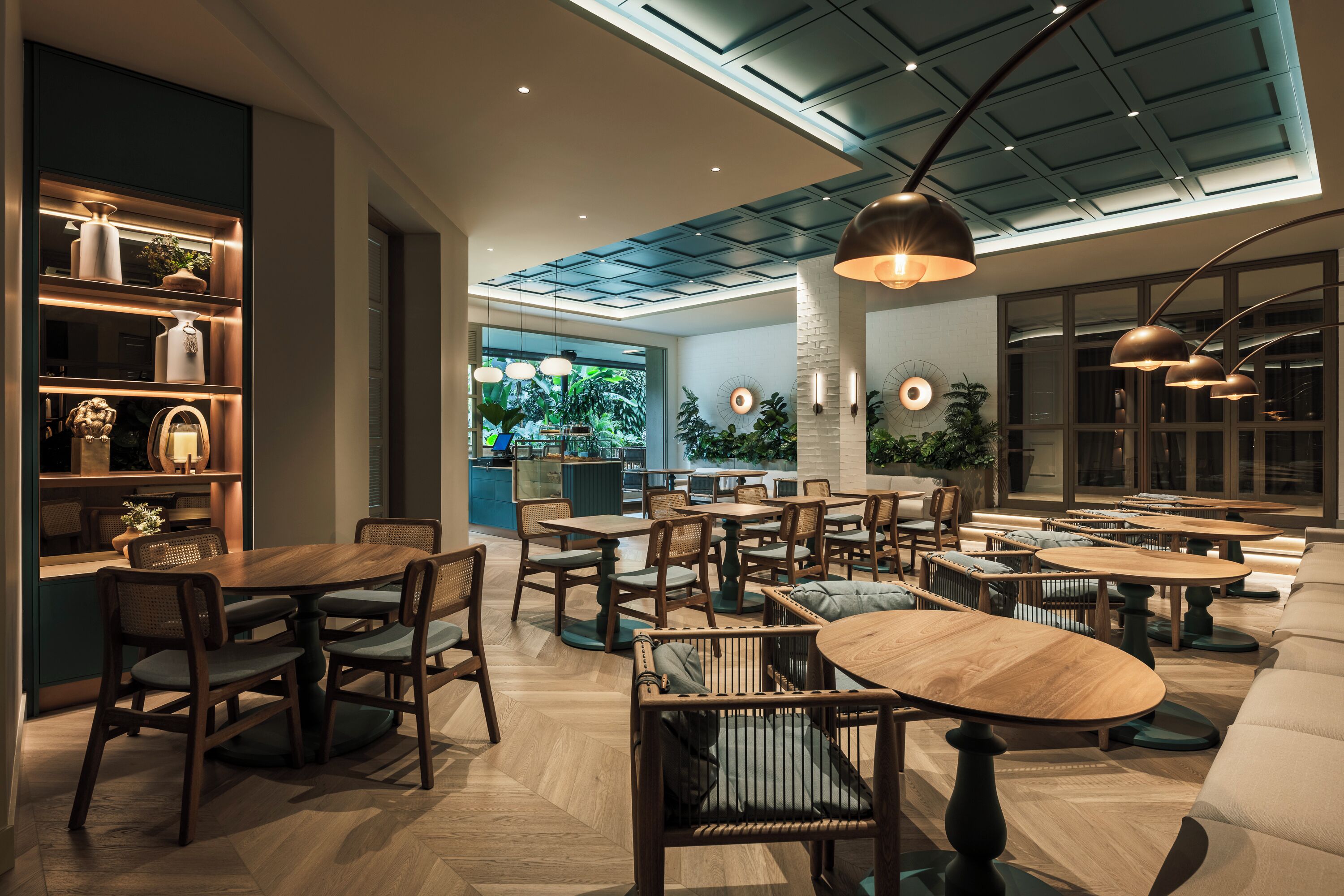A modern restaurant interior with wooden tables and chairs, a central seating area with a sofa, and a ceiling with a geometric pattern.