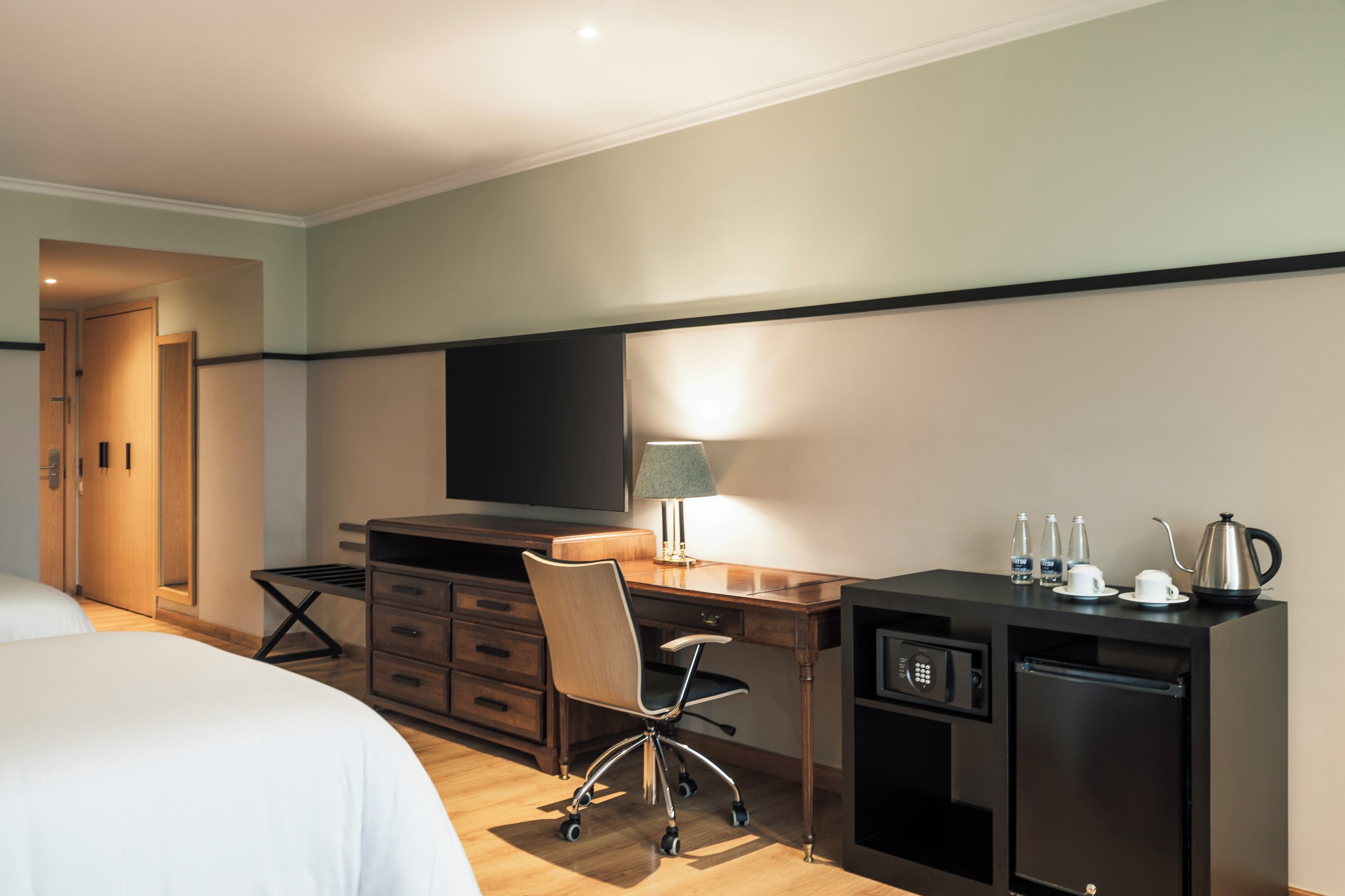 Habitación de hotel con cama, escritorio con silla, televisor, lámpara, cafetera y minibar.