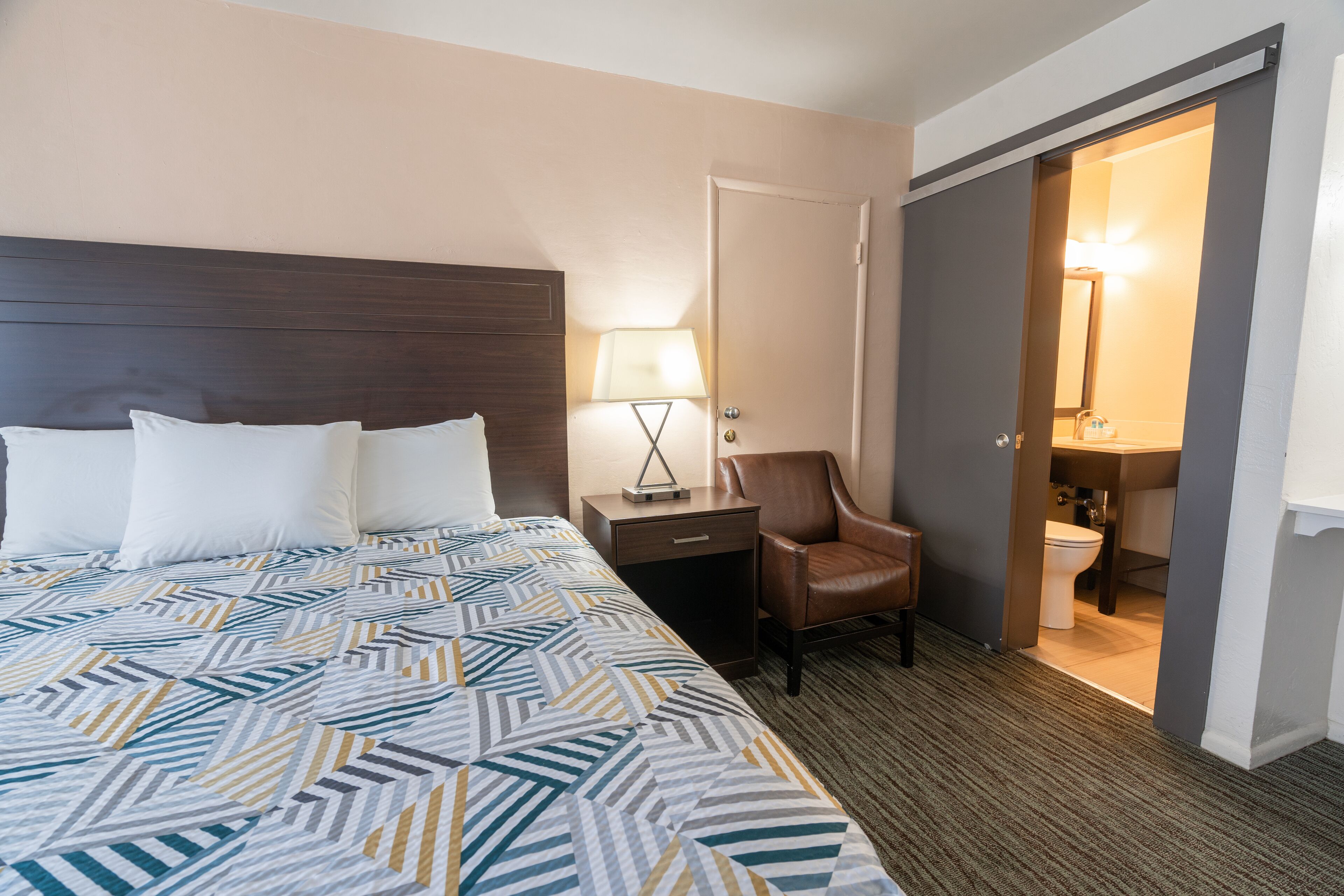 Een hotelkamer met een groot bed, een nachtkastje met een lamp, een stoel en een badkamer zichtbaar door een geopende deur.