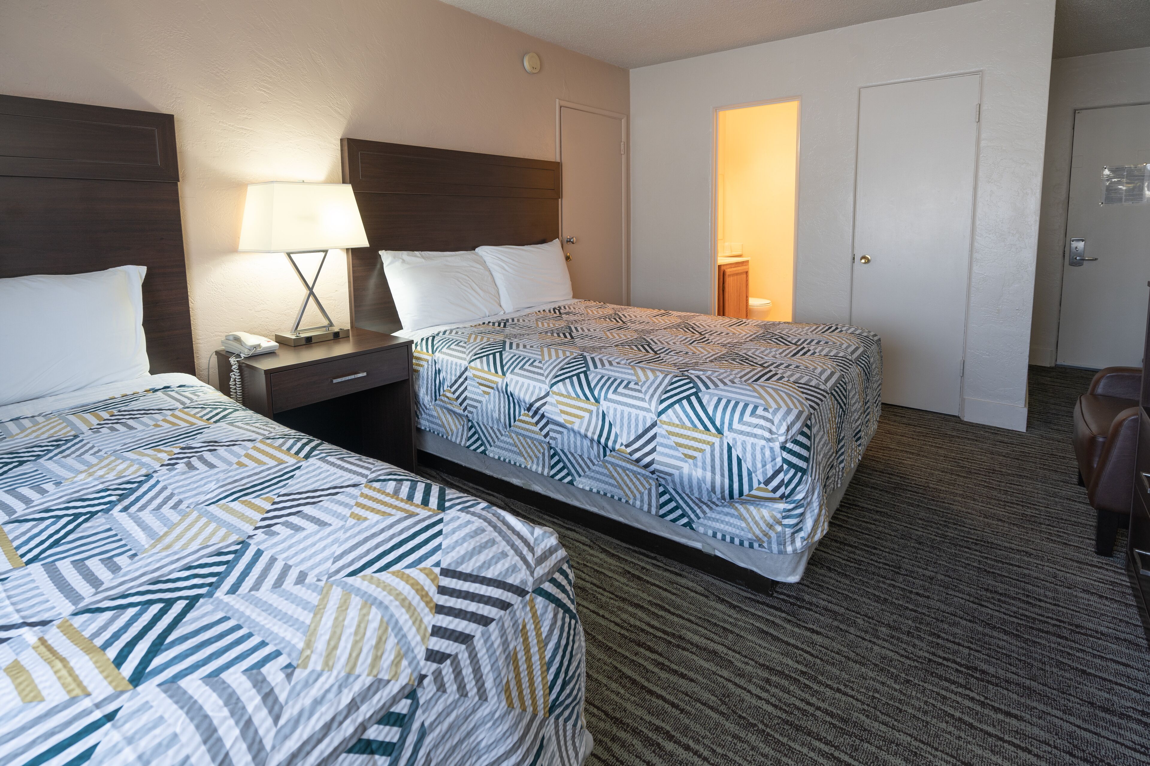 Habitación de hotel con dos camas, una mesita de noche con lámpara y una puerta que da a un baño.