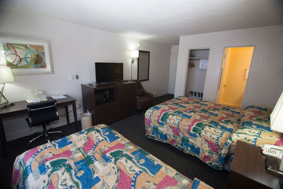 A hotel room with two beds, a desk with a chair, a TV, a desk lamp, a trash can, and a door leading to another room.