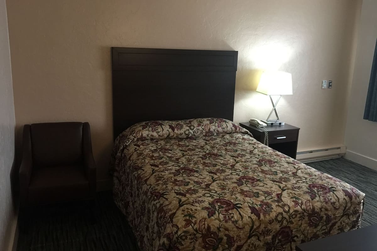 A hotel room with a bed, a chair, a nightstand with a lamp, and a wall-mounted phone.