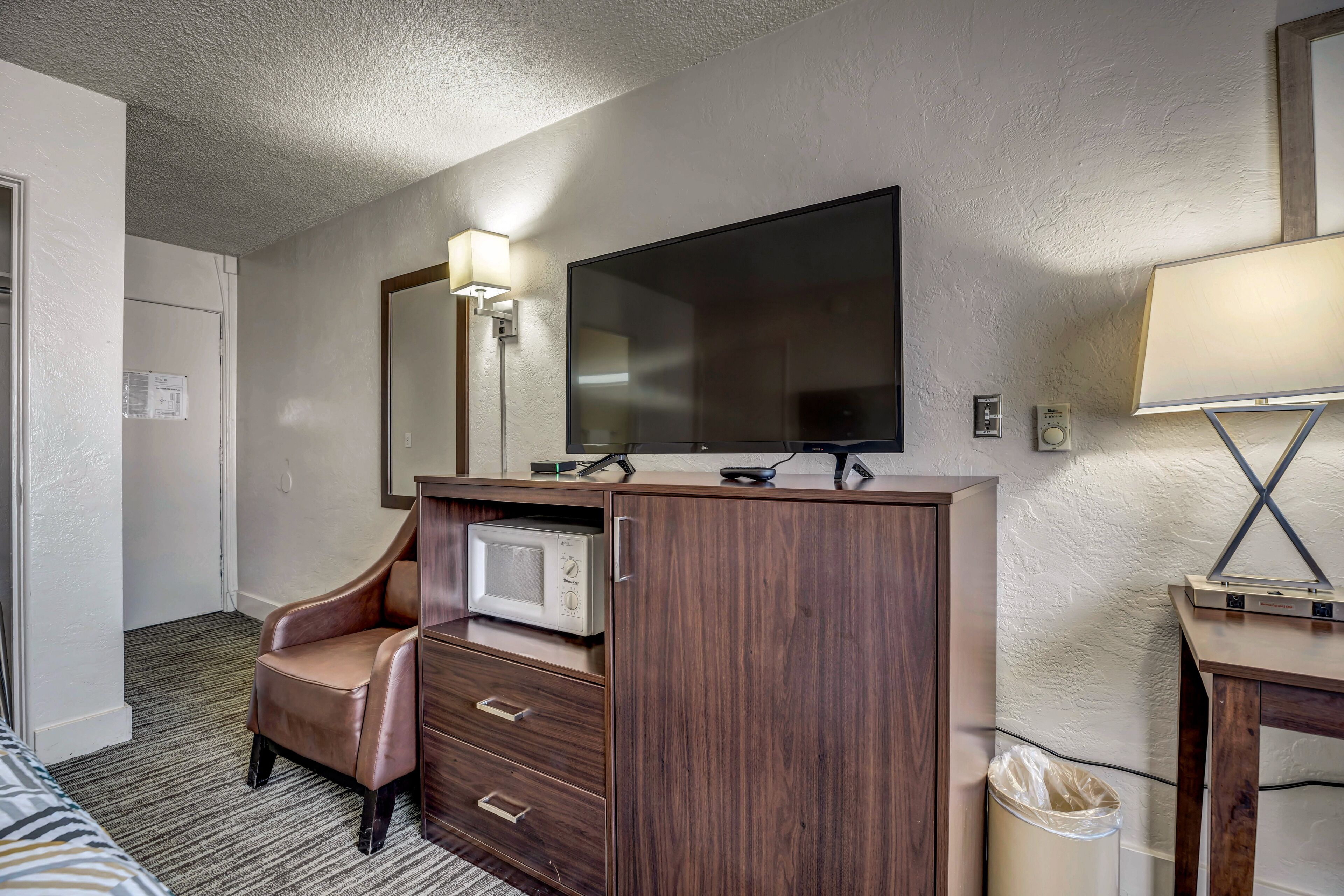 A hotel room with a wooden desk, a television, a microwave, a chair, and a trash can.