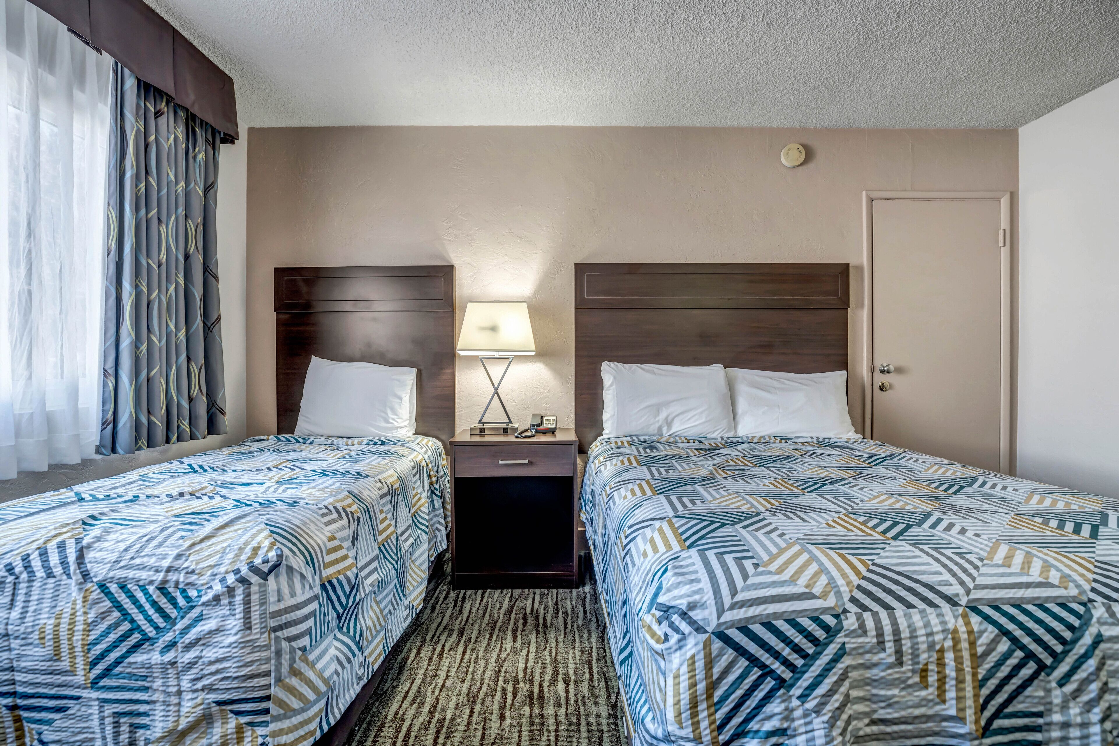 A hotel room with two beds, a nightstand with a lamp, and a door.