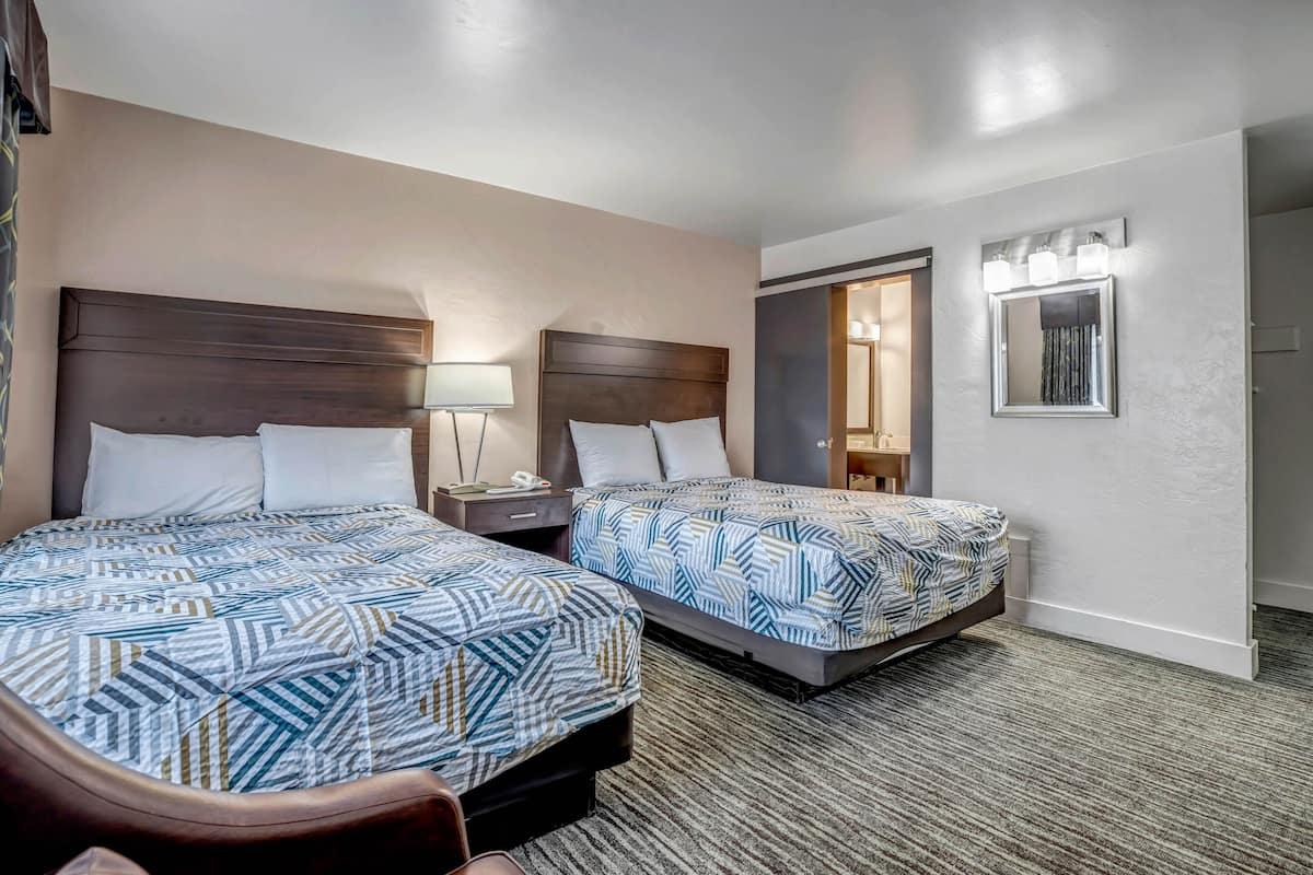 A hotel room with two beds, a nightstand, a lamp, a mirror, and a door leading to another room.
