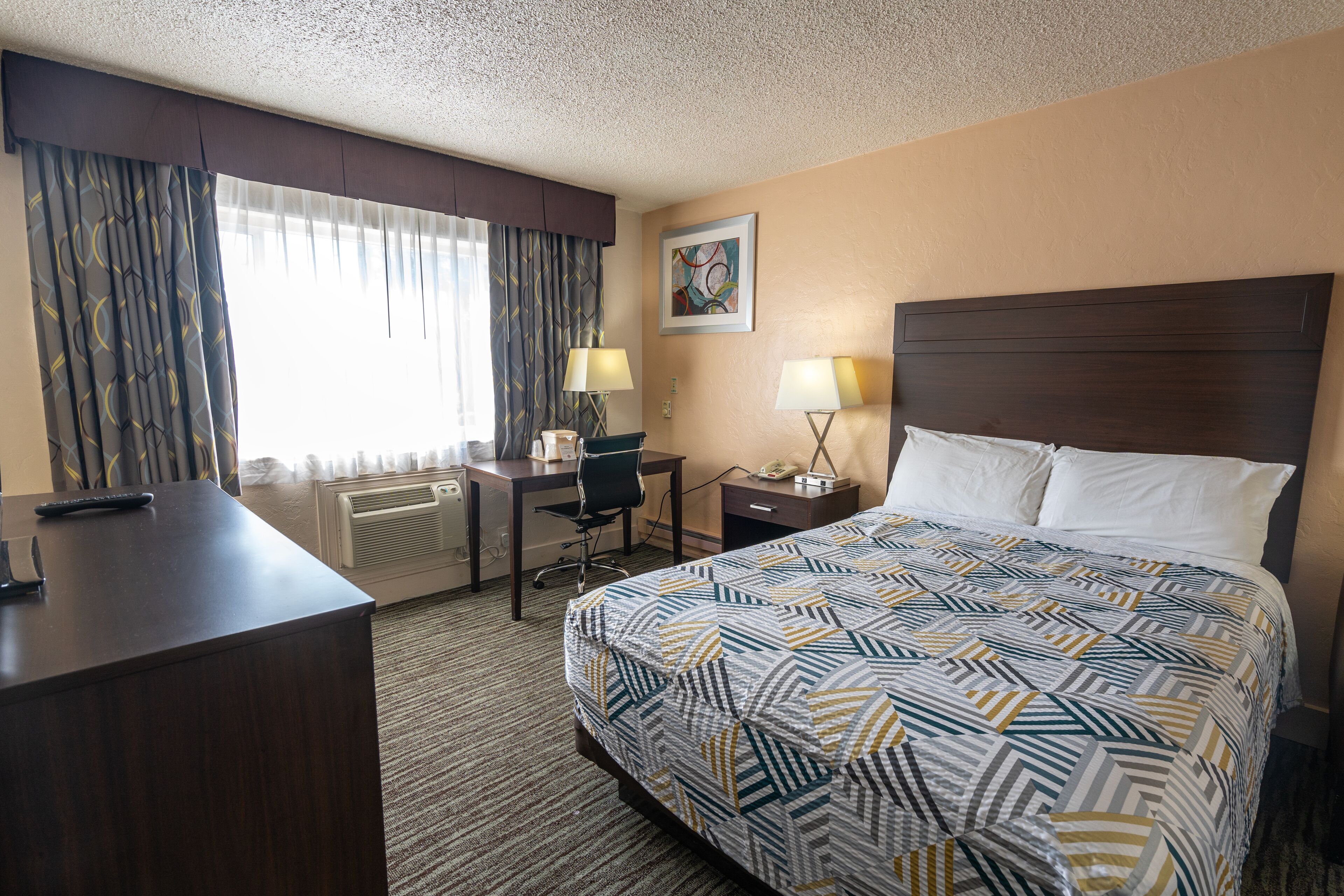 A hotel room with a bed, desk, chair, two bedside lamps, a painting on the wall, and a window with curtains.