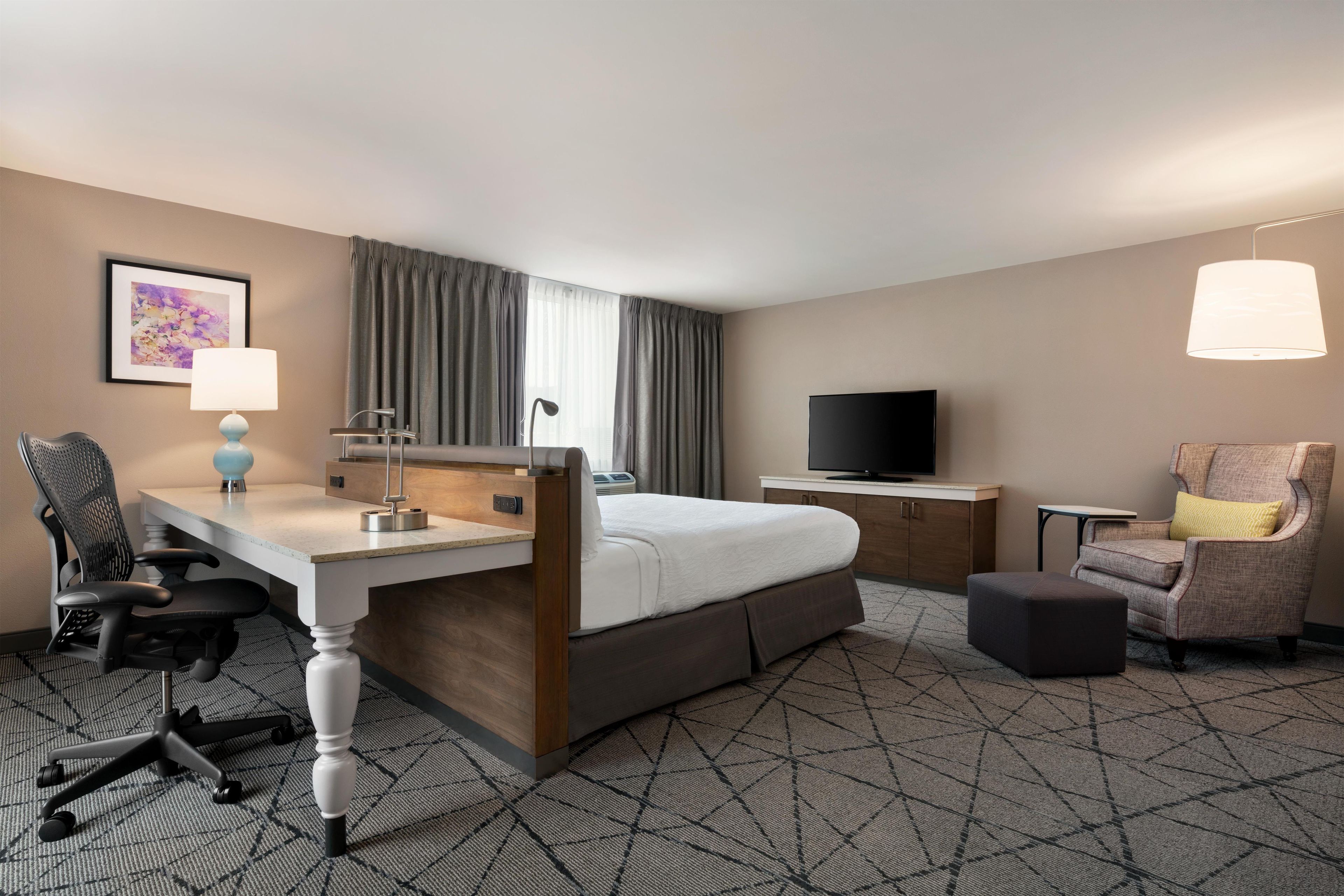 A hotel room with a bed, desk, chair, television, and a lamp.