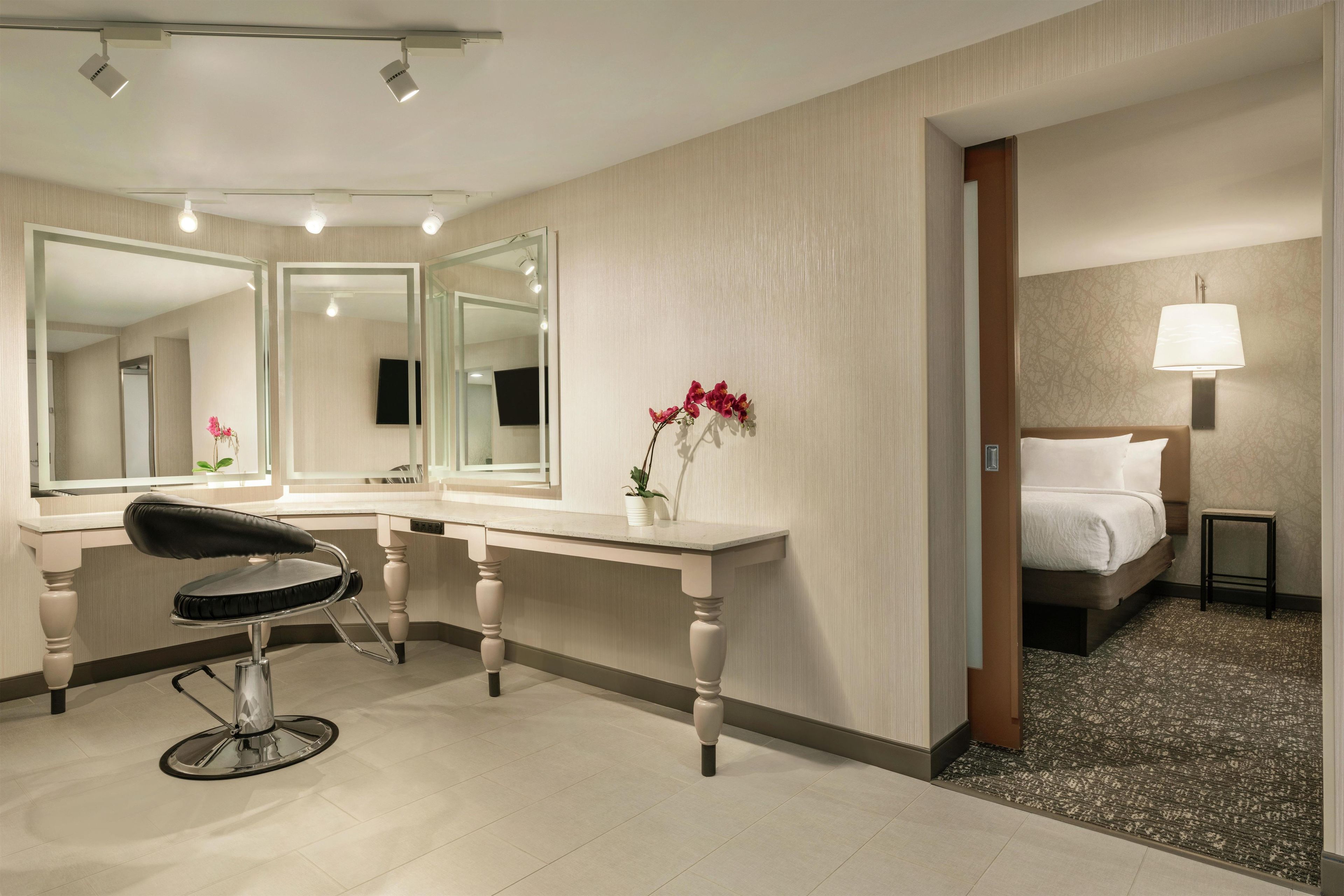 A hotel room with a large mirror, a desk, a chair, a bed, and a nightstand with a lamp.