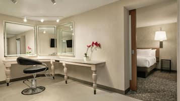 A hotel room with a large mirror, a desk, a chair, a bed, and a nightstand with a lamp.