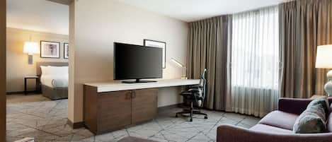 Habitación de hotel con cama, escritorio con televisor, silla, sofá y ventana grande con cortinas.