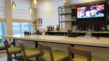 A bar area with a long counter, multiple bar stools, a television screen, and shelves with bottles and glasses.