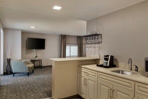 Habitación de hotel con televisor de pantalla plana, una pequeña zona de estar con una silla y una mesa, una kitchenette con cafetera y un ventanal grande.