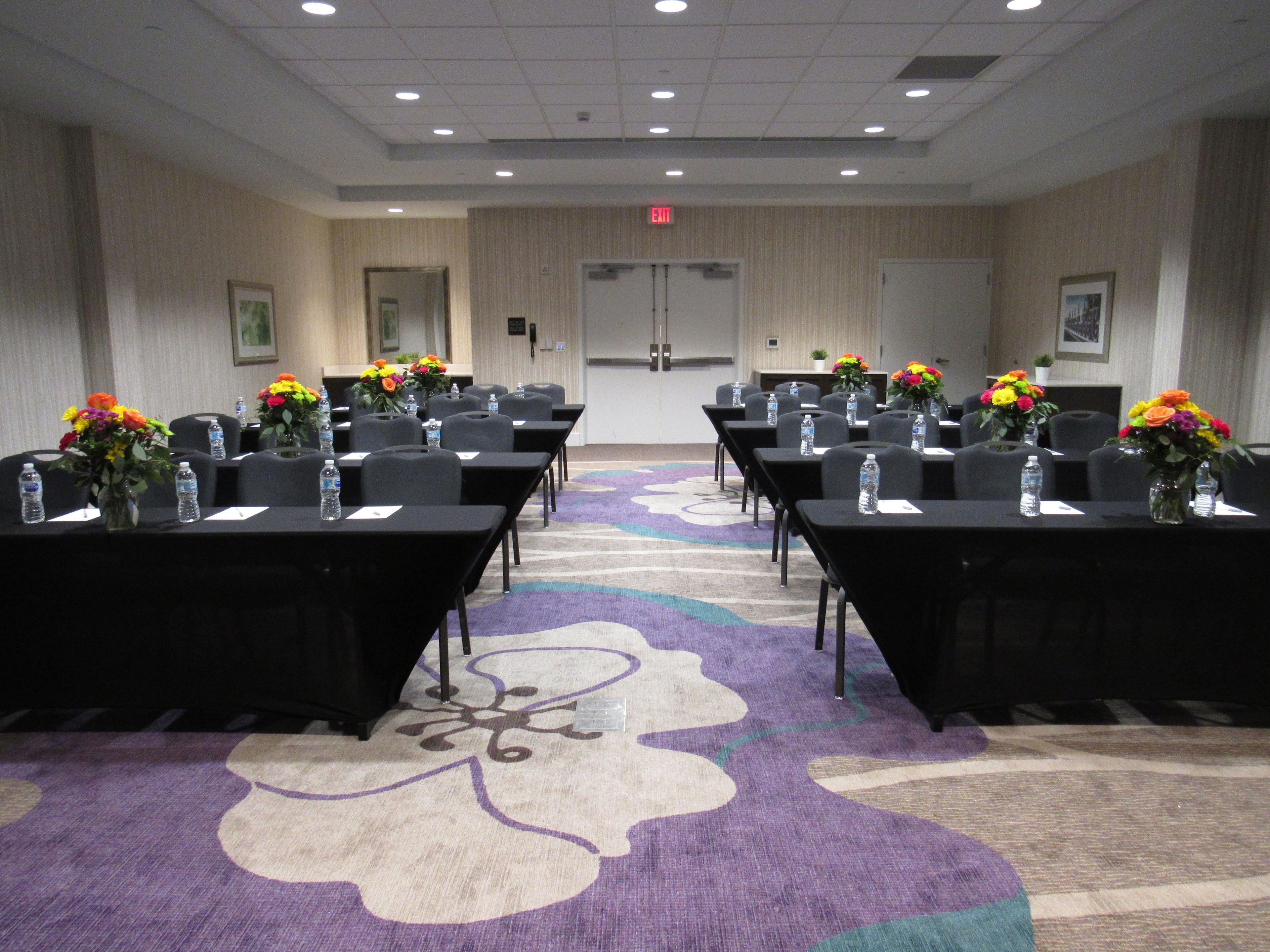 Une salle de conférence avec des rangées de tables, de chaises et des compositions florales en guise de décoration, ainsi qu’un tapis orné de motifs floraux et une porte sur laquelle est apposée une signalétique indiquant la sortie.