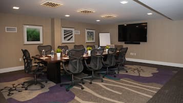 A conference room with a long table, multiple chairs, a whiteboard, and a flat-screen TV mounted on the wall.