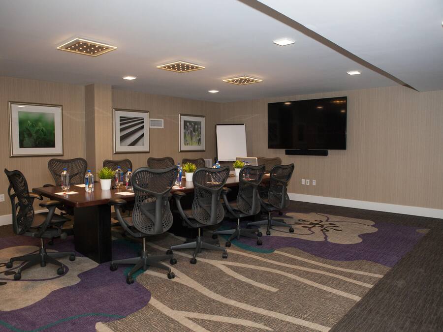 A conference room with a long table, multiple chairs, a whiteboard, and a flat-screen TV mounted on the wall.