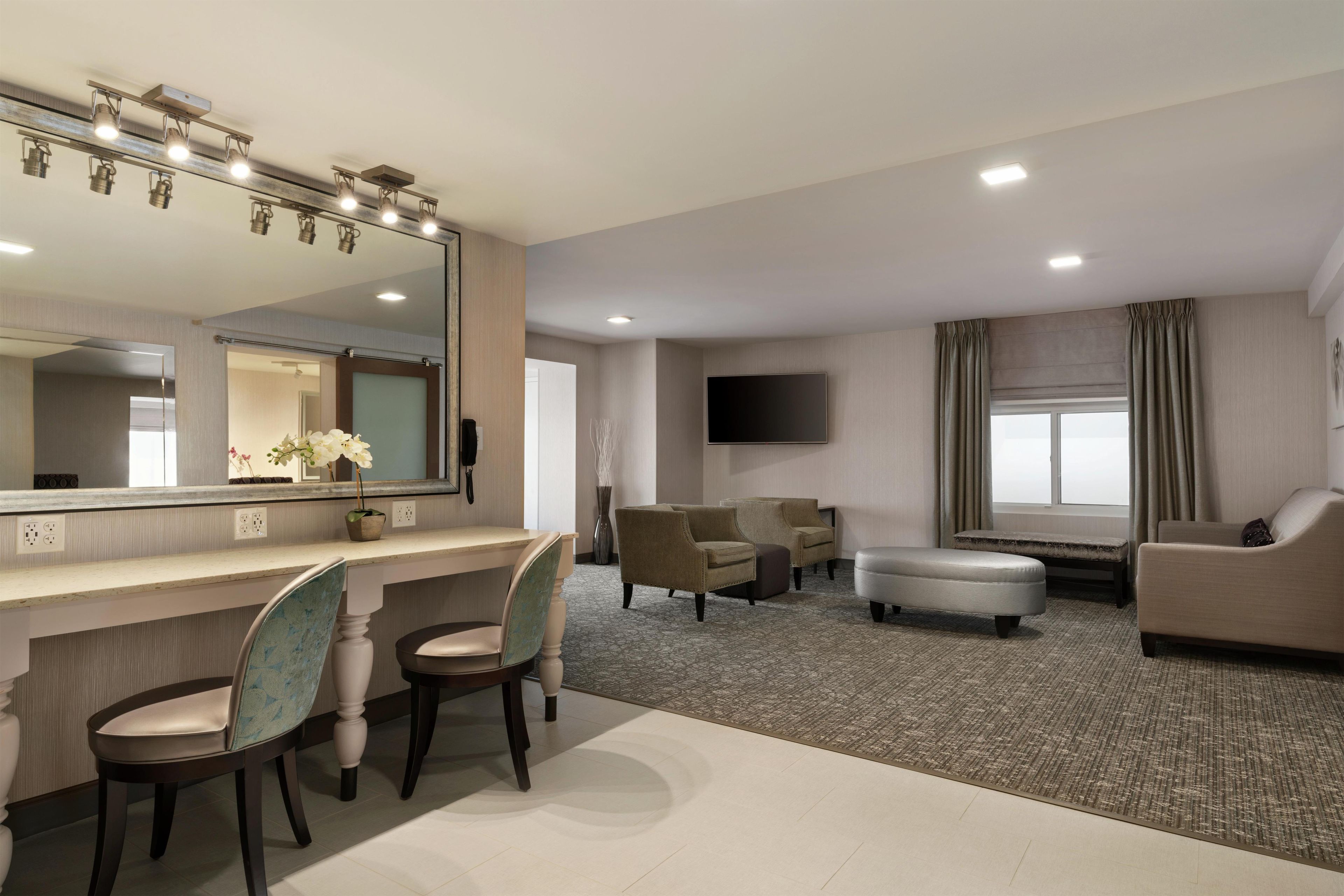 A hotel room with a vanity, two chairs, a sofa, a flat-screen TV, and a large mirror.