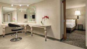 A hotel room with a large mirror, a vanity with a chair, a bed with a white comforter, and a small bedside table with a lamp.