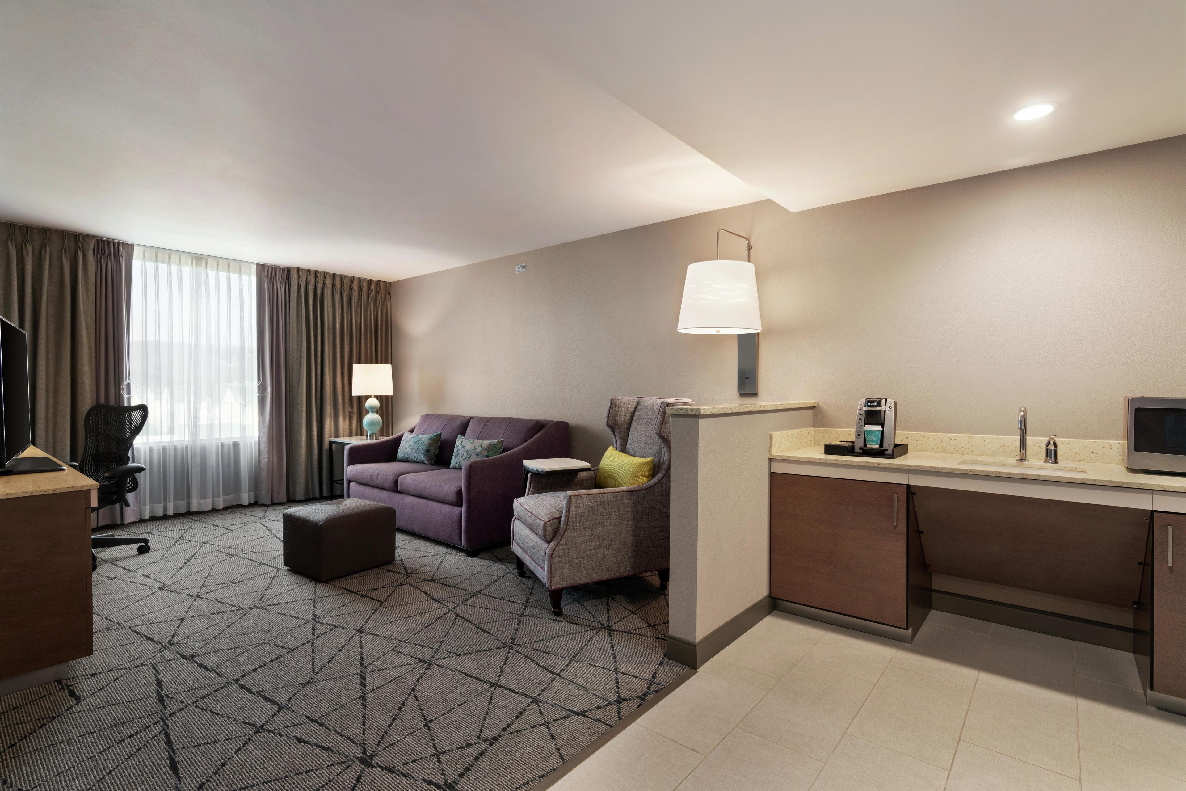 A hotel room with a sofa, armchair, desk, and a coffee machine.