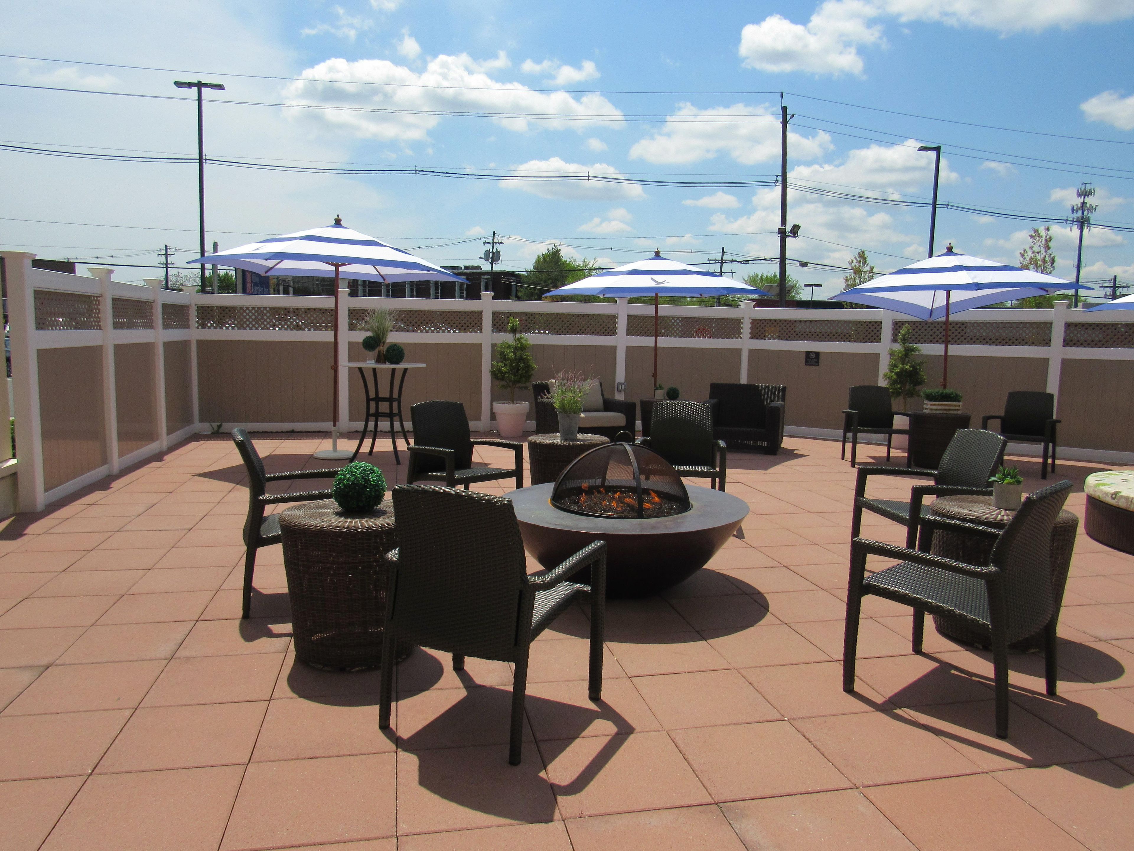 Ein Terrassenbereich im Freien mit Fliesenböden, Korbmöbeln, einer Feuerstelle und Sonnenschirmen, die Schatten spenden.
