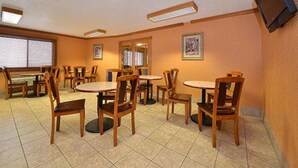 A dining area with multiple tables and chairs, a television mounted on the wall, and windows with blinds.