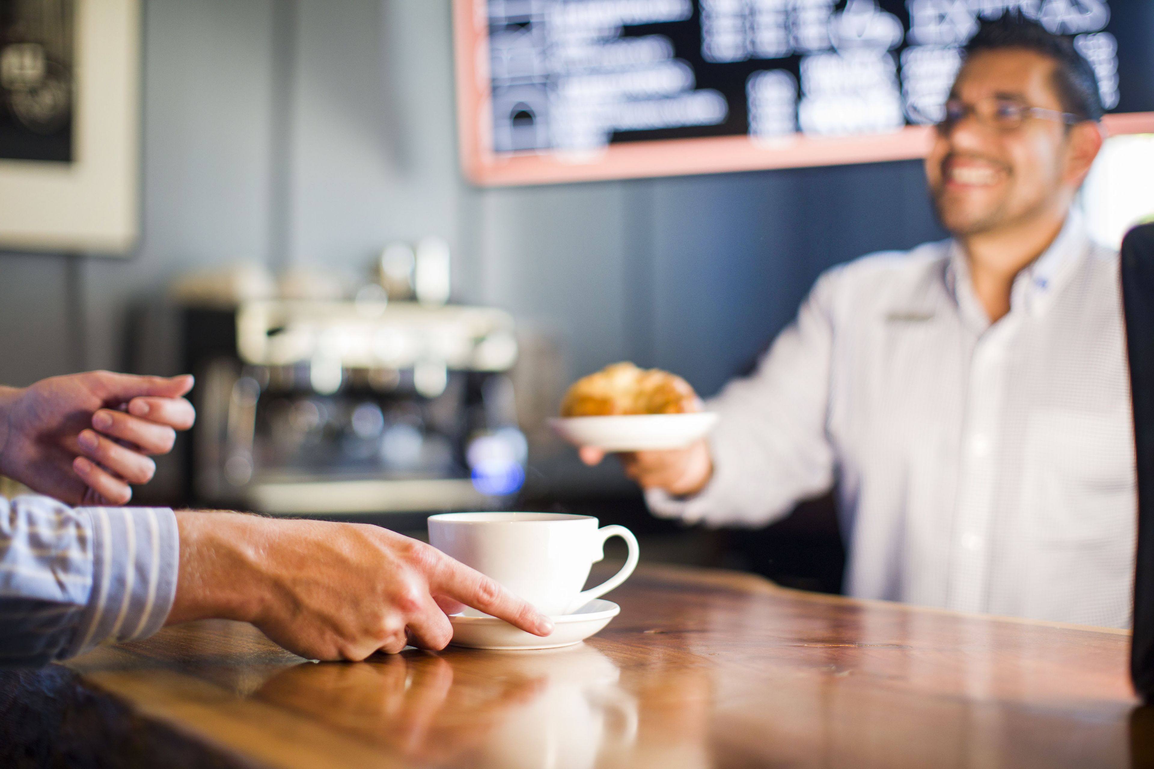 A person holding a plate with a croissant, standing at a counter with a cup and saucer.