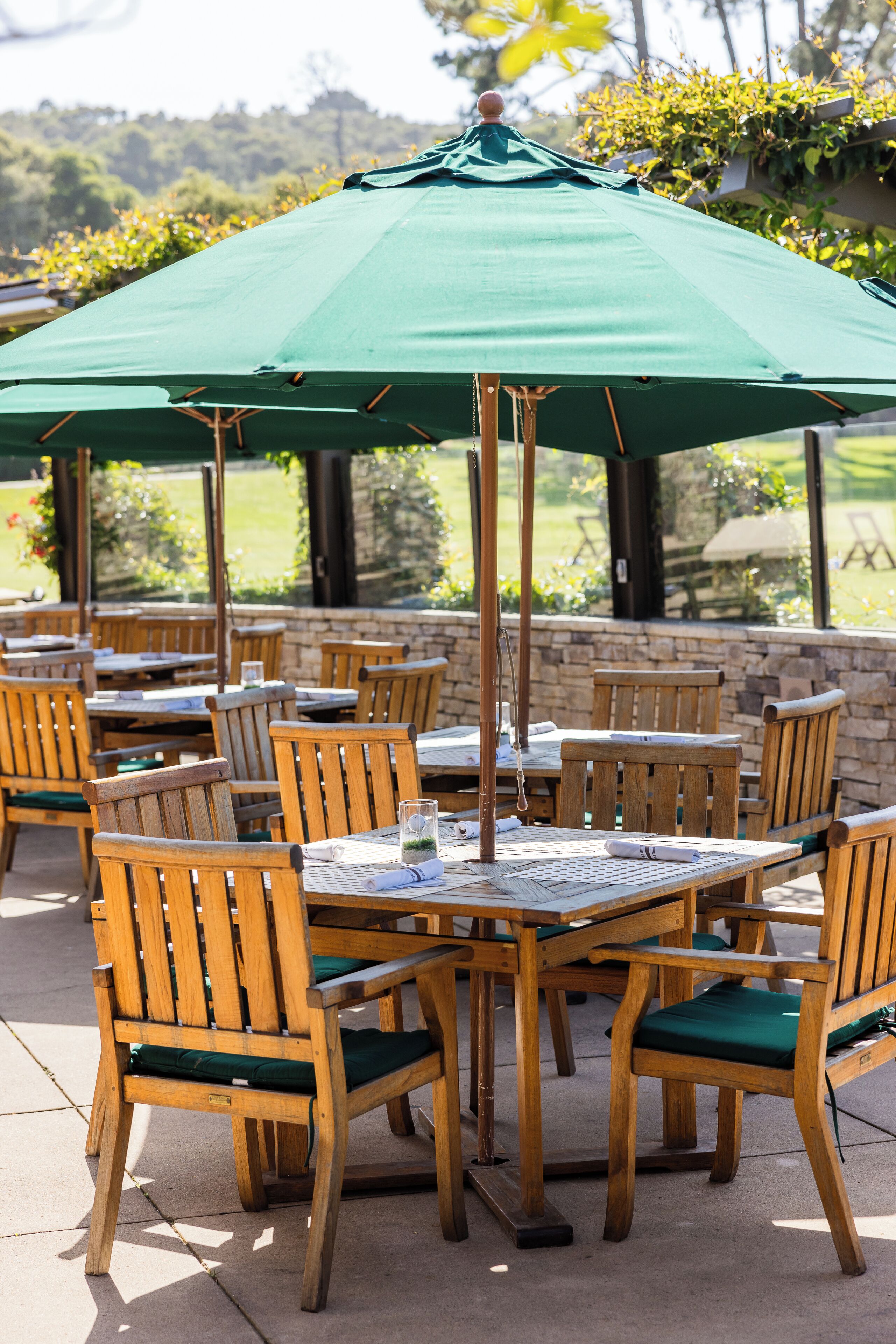 An outdoor patio area with wooden tables and chairs, a green umbrella, and a stone wall.