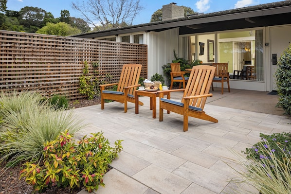 Une terrasse extérieure avec des chaises et une table en bois, entourée de plantes et d’une clôture en bois.