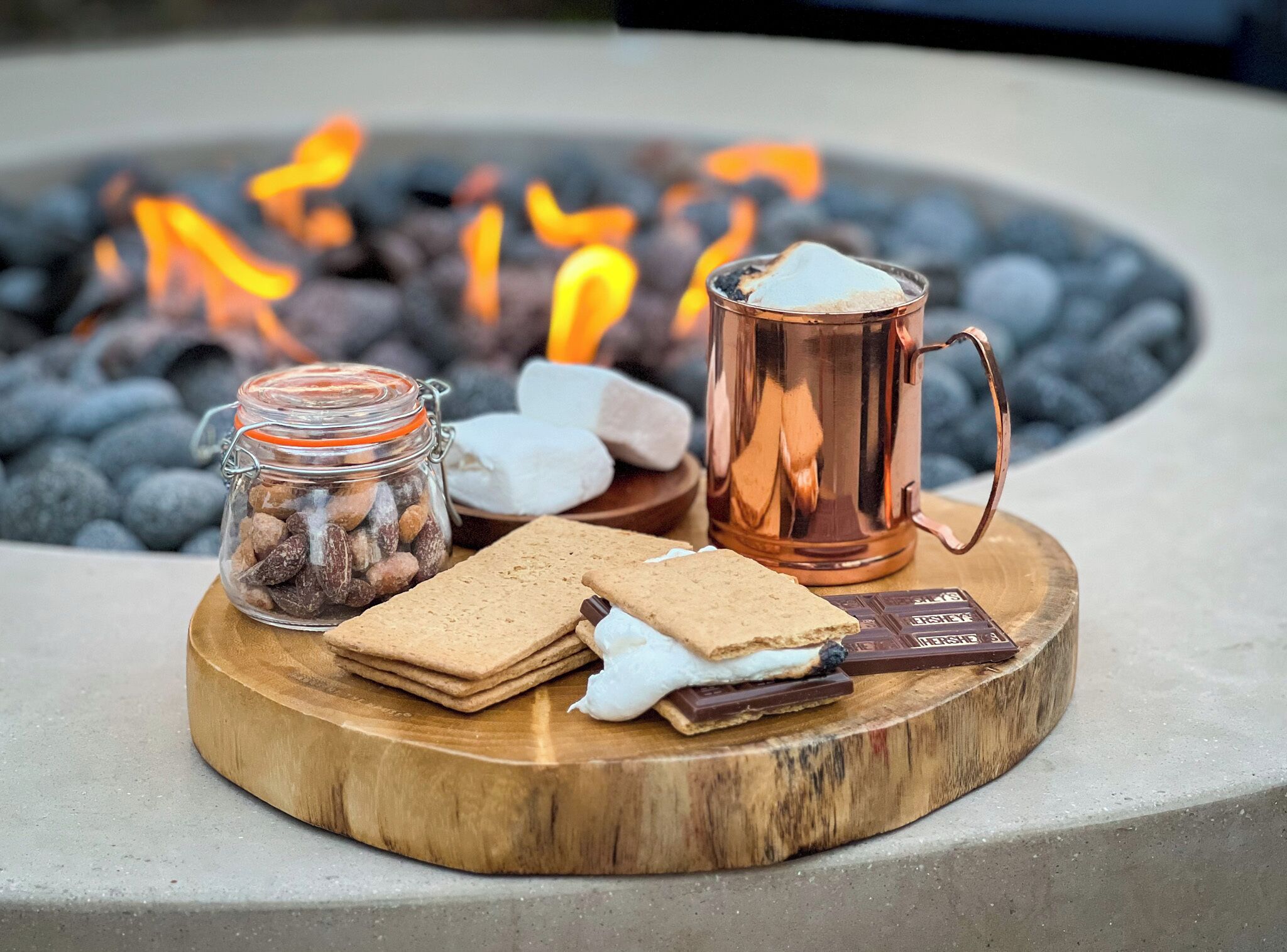A wooden tray with snacks and a mug, set against a backdrop of a fire pit.