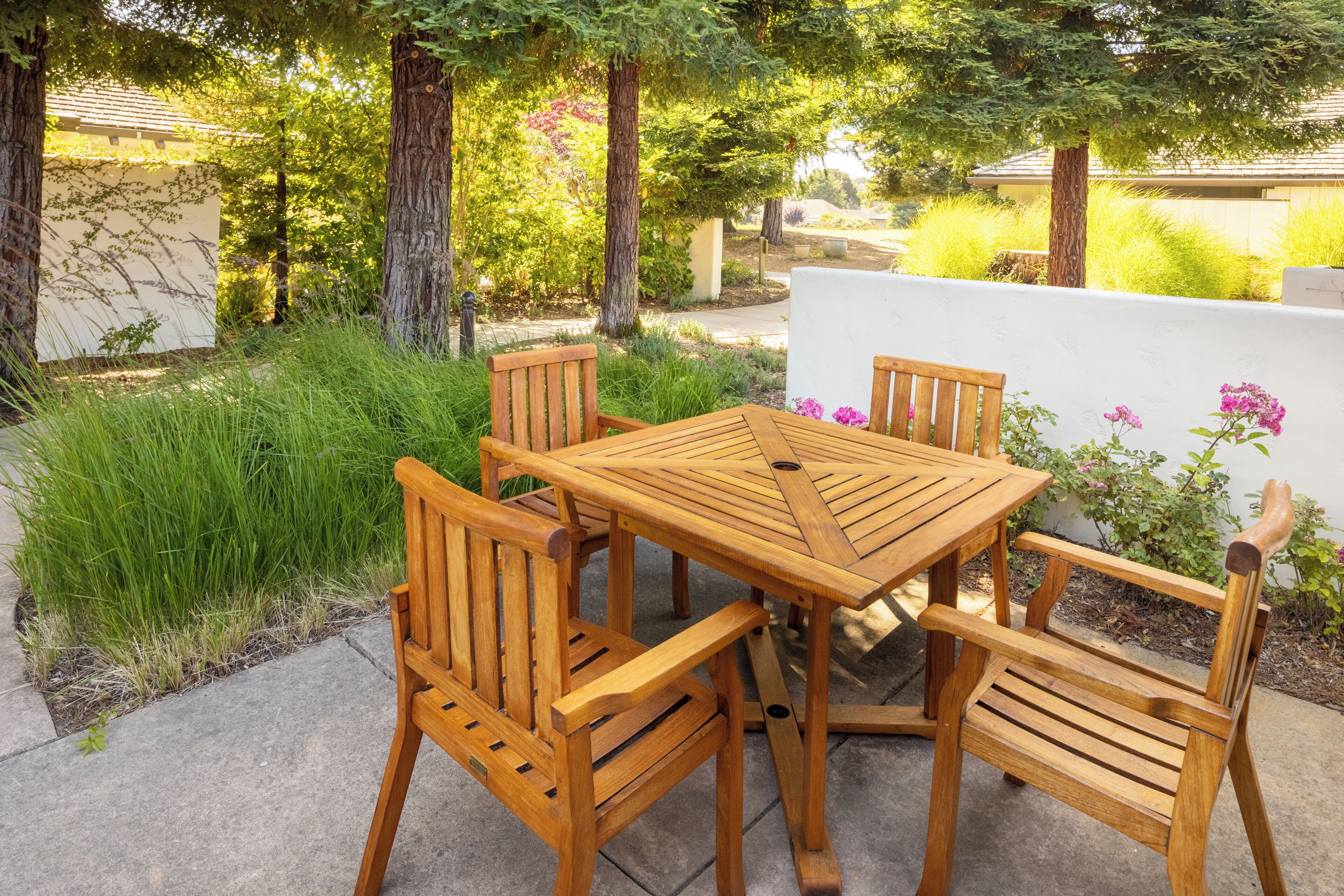 A wooden outdoor dining set with four chairs and a table, surrounded by greenery and a white wall.