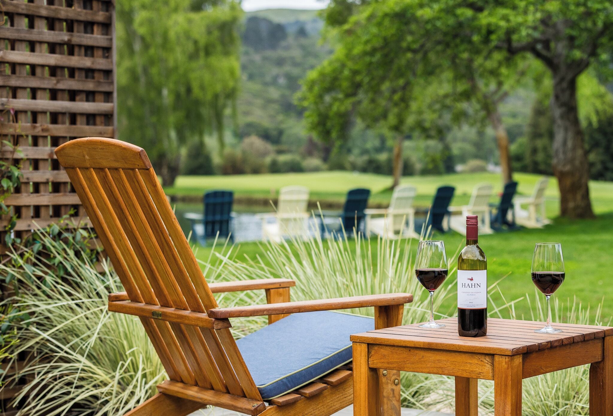 A wooden chair with a cushion, a wooden table with two wine glasses and a bottle, and a view of a grassy area with trees.