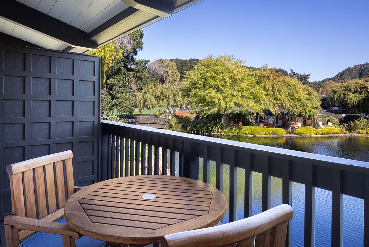 A balcony with a wooden table and chairs, overlooking a body of water and a landscaped garden.
