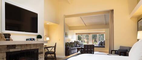 A bedroom with a stone fireplace, a large bed, a TV mounted above the fireplace, and a seating area with a view of the outdoors.