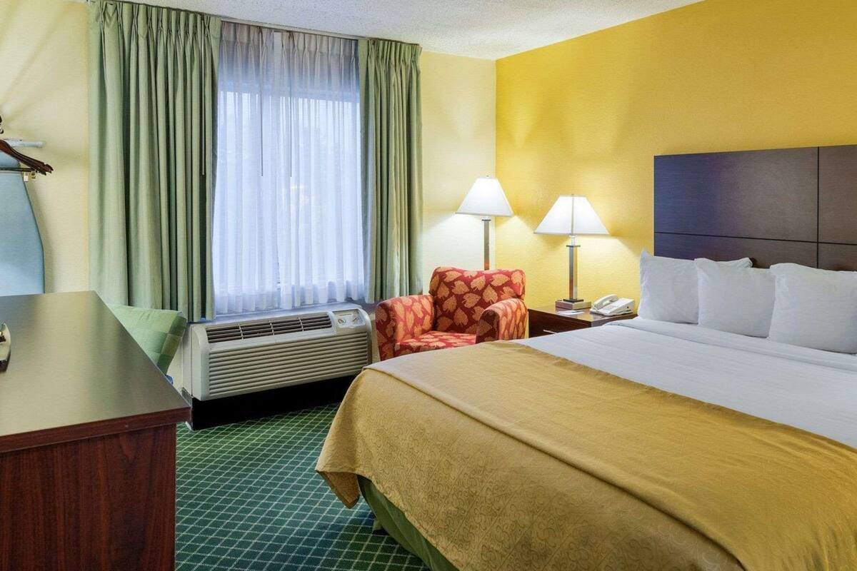 Una habitación de hotel con cama, dos lámparas de noche, una silla, escritorio y ventana con cortinas.