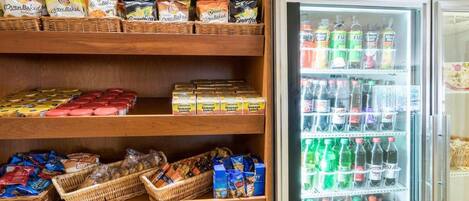 Zona de exhibición de bocadillos y bebidas con estantes llenos de diferentes opciones y un refrigerador con bebidas embotelladas.