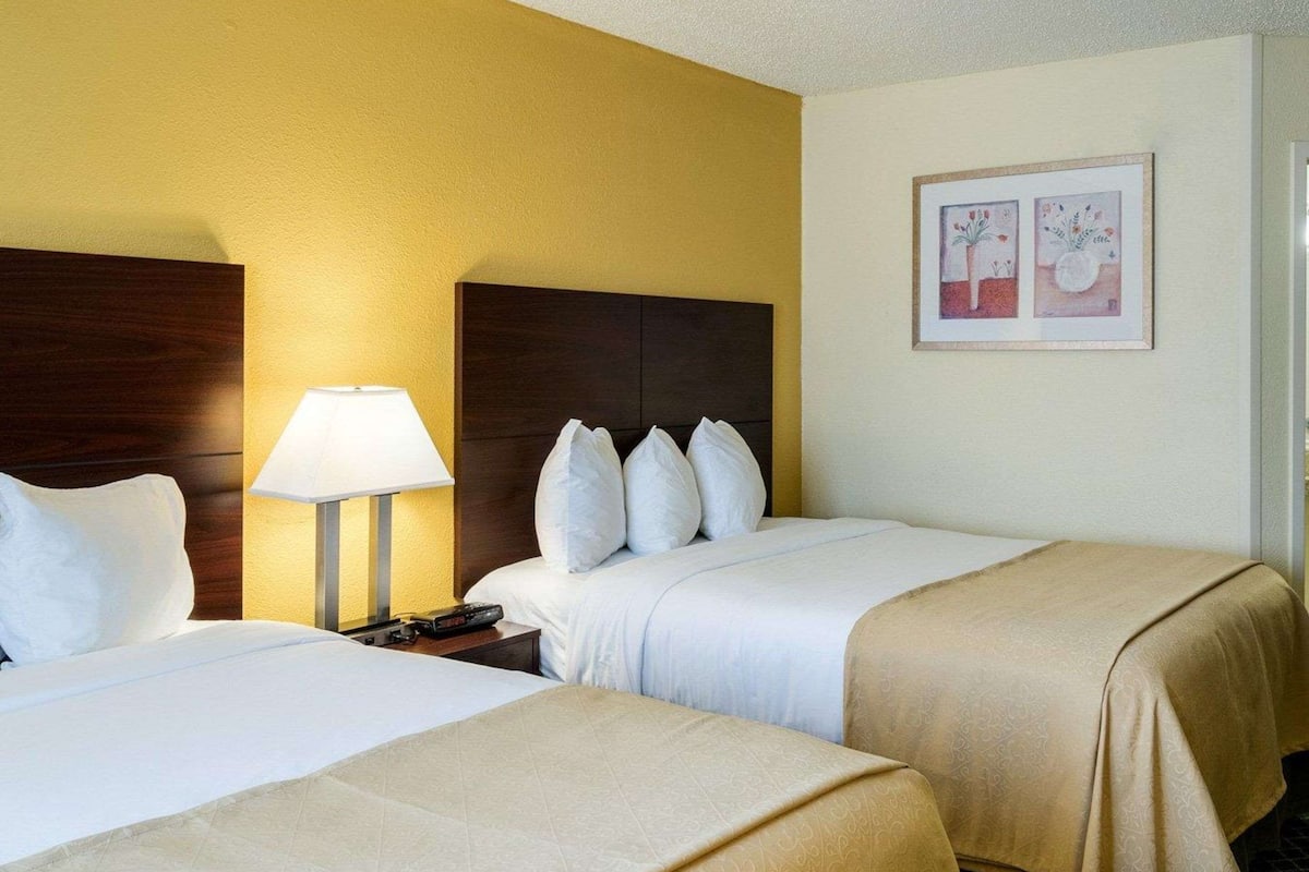 Una habitación de hotel con dos camas, una mesita de noche con lámpara y dos cuadros en la pared.