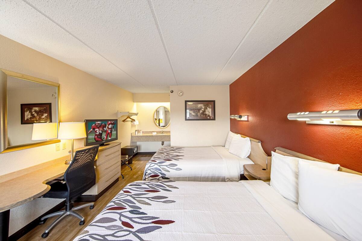 A hotel room with two beds, a desk, a chair, a TV, a mirror, and a wall-mounted lamp.