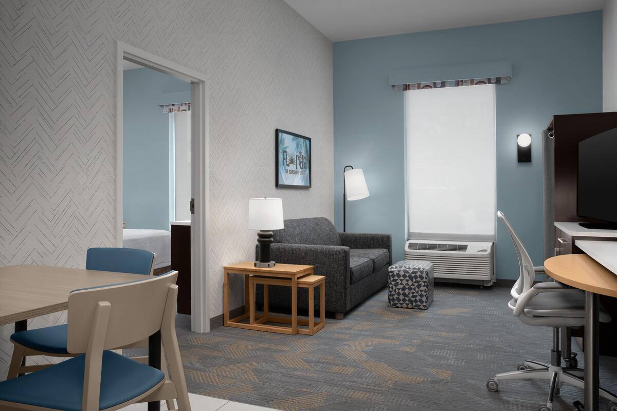 A hotel room with a herringbone-patterned wall, a gray sofa, a wooden side table with a lamp, a gray patterned rug, a small ottoman, a desk with a chair, and a television.
