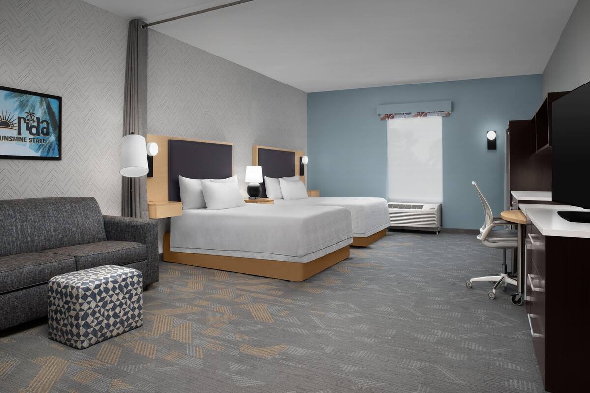 A hotel room with a herringbone-patterned carpet, two beds with white linens, a grey sofa, a patterned ottoman, a desk with a chair, and a large window with a view.