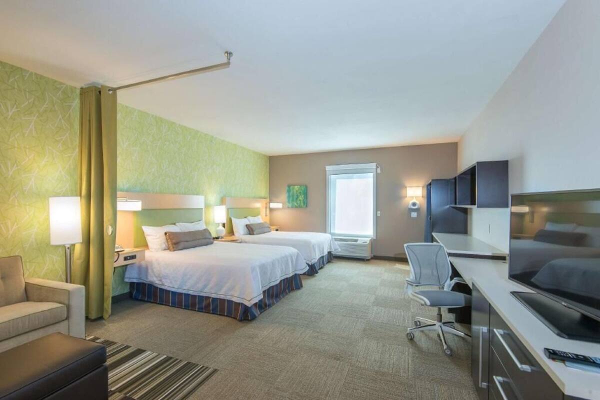 A hotel room with two beds, a desk with a TV, a chair, a lamp, and a window.