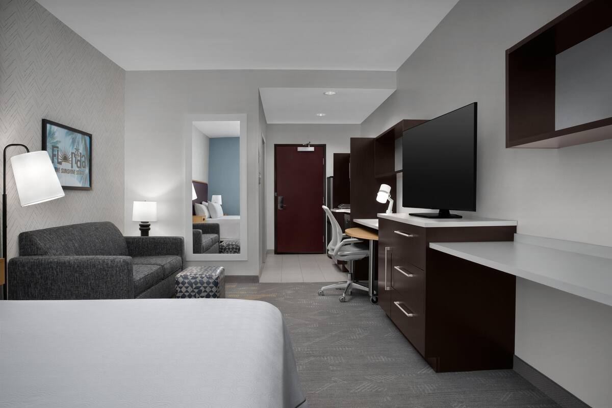A modern hotel room with a bed, a desk with a computer, a TV, and a lamp.