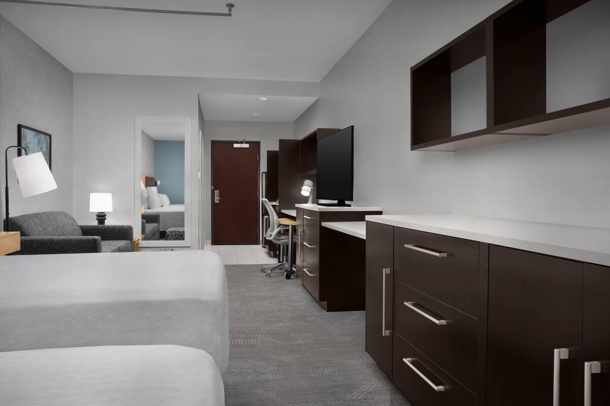 A modern hotel room with a large bed, a desk, a chair, a TV, and a wardrobe.