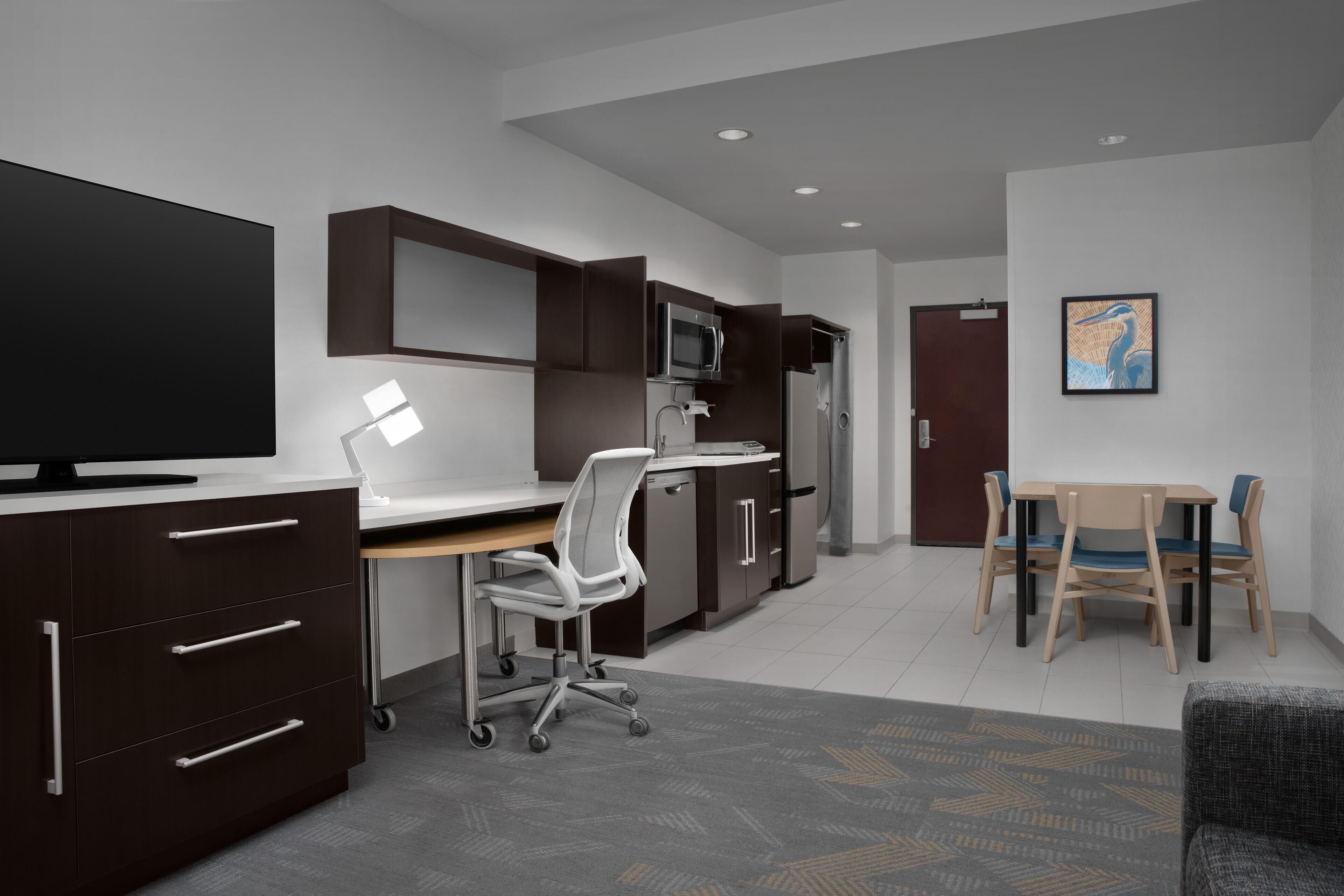 A modern kitchen with a flat-screen TV, microwave, and refrigerator. There is a dining area with a table and chairs.