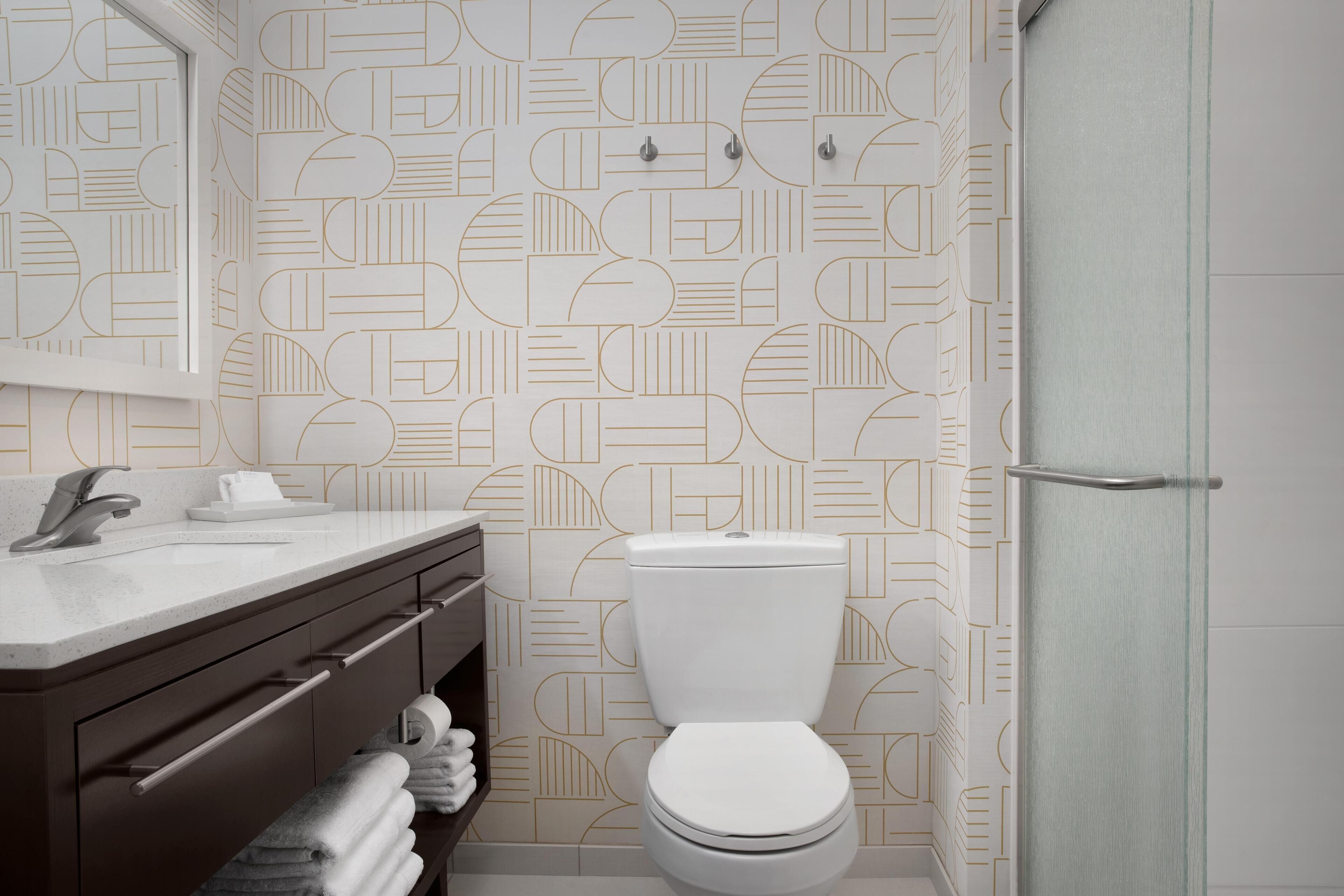 A modern bathroom with a geometric patterned wall, a white toilet, a dark wooden vanity with a mirror, and a glass shower enclosure.