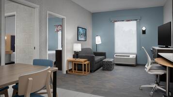 A hotel room with a dining table, chairs, a sofa, a TV, and a window with curtains.
