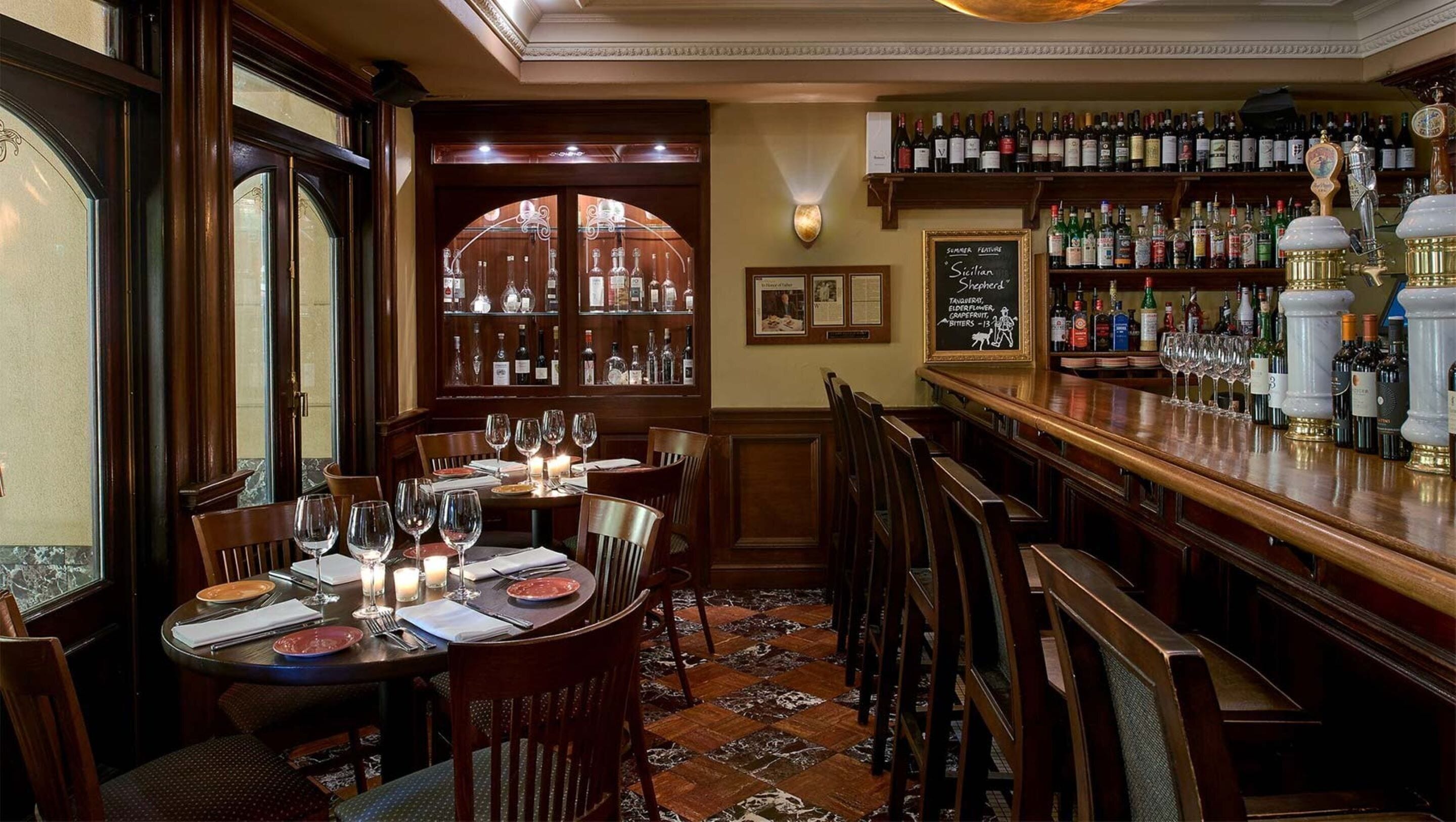 A bar area with a wooden counter, shelves of liquor bottles, and a table set for dining with chairs and wine glasses.