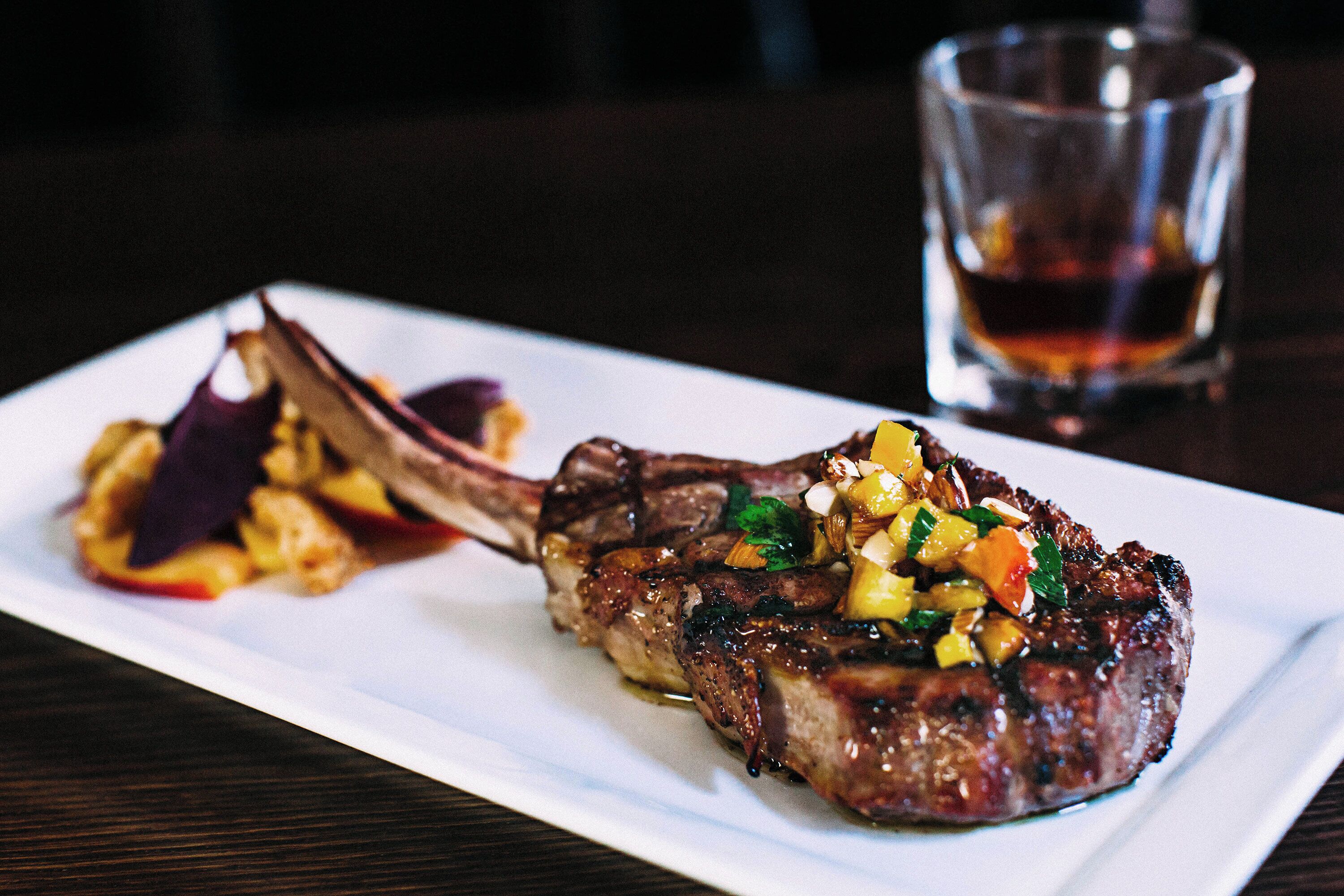 A plate with a grilled meat dish, a glass of beverage, and a dark wooden table.