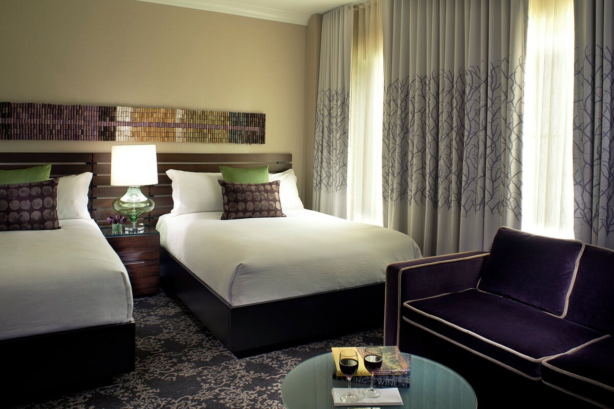 A hotel room with two beds, a nightstand with a lamp, a chair, and a small table with a book and glasses.
