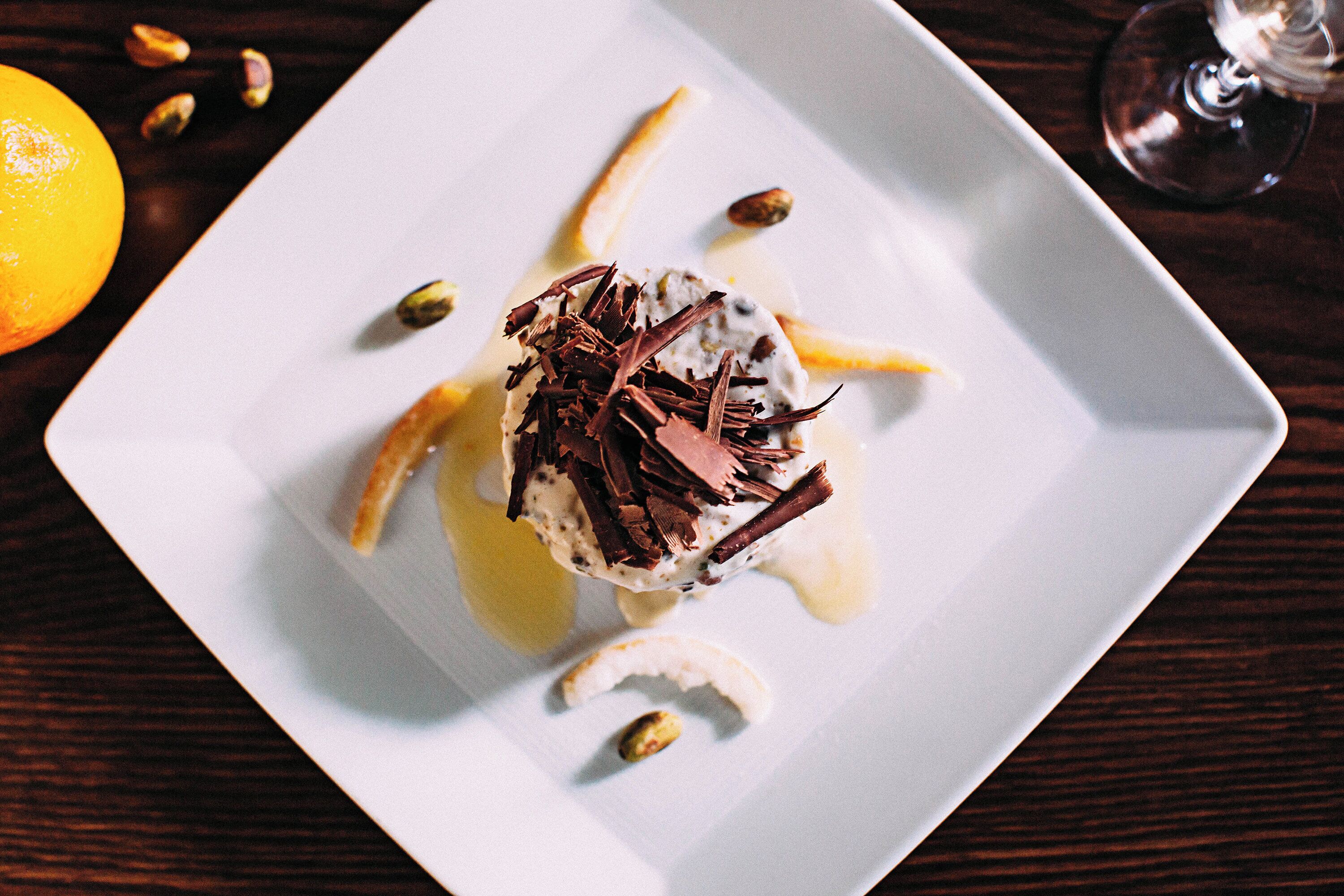 A dessert on a square white plate with chocolate shavings and pistachios.