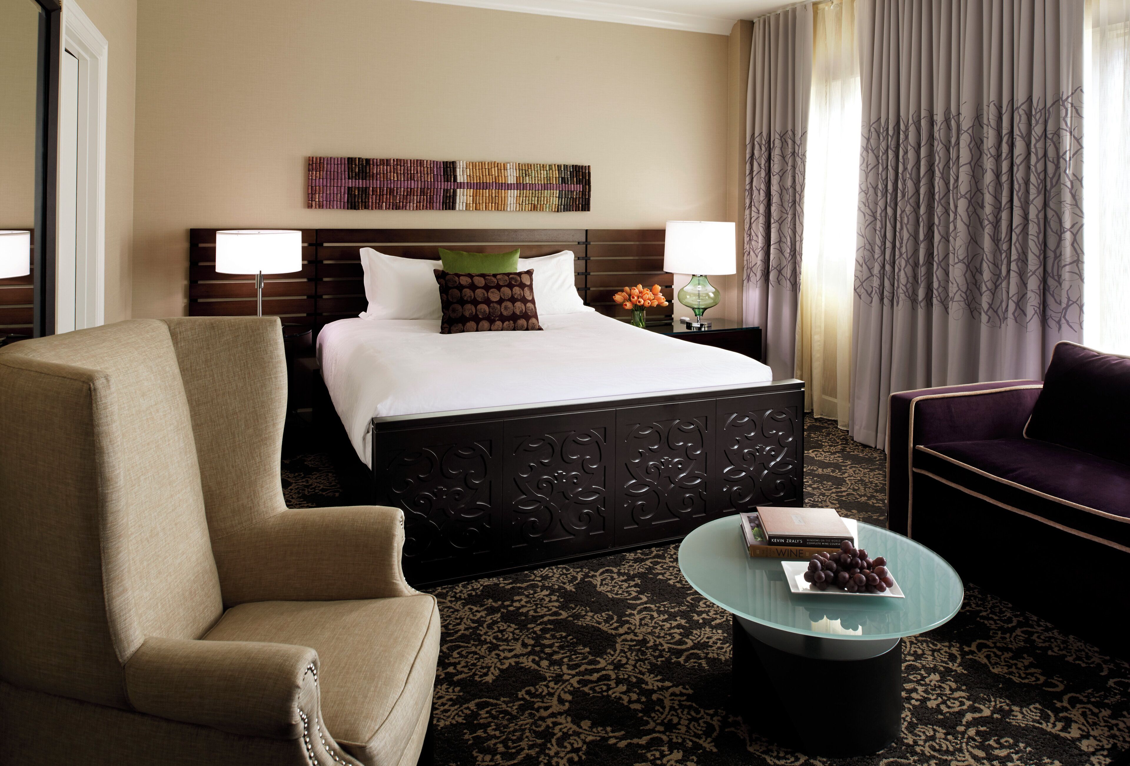 A hotel room with a large bed, two bedside lamps, a green vase with flowers, a glass coffee table with a book and grapes, and a dark patterned carpet.